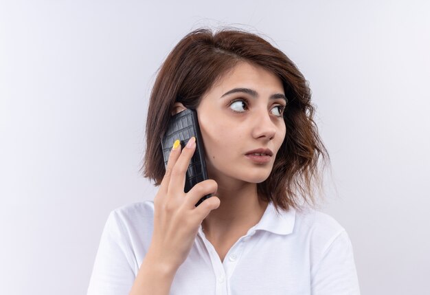 Jeune fille aux cheveux courts portant un polo blanc à côté confus tout en parlant au téléphone mobile