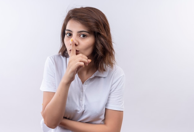 Jeune fille aux cheveux courts portant un polo blanc faisant un geste de silence avec fingr sur les lèvres