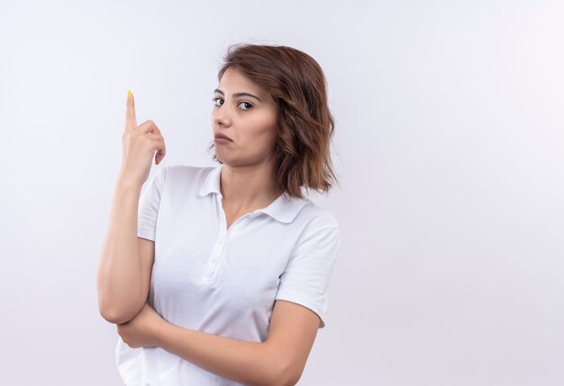 Jeune fille aux cheveux courts portant un polo blanc à la recherche de pointage suspect avec l'index vers le haut