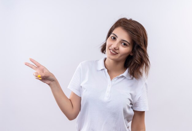 Jeune fille aux cheveux courts portant un polo blanc souriant joyeusement montrant le signe de la victoire