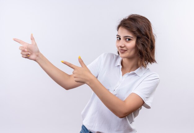 Jeune fille aux cheveux courts portant un polo blanc souriant pointant avec l'index sur le côté