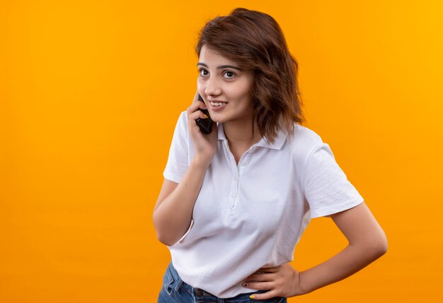 Jeune fille aux cheveux courts portant un polo blanc souriant avec un visage heureux tout en parlant au téléphone mobile