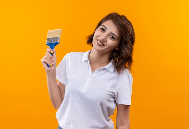 Jeune fille aux cheveux courts portant un polo blanc tenant un pinceau souriant joyeusement