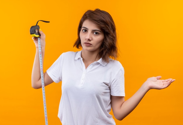 Jeune fille aux cheveux courts portant un polo blanc tenant un ruban à mesurer regardant la caméra confus