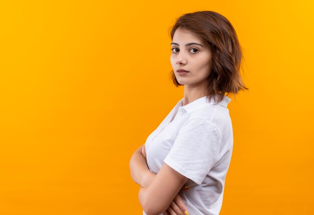 Jeune fille aux cheveux courts portant un polo debout sur le côté avec les bras croisés regardant la caméra avec un visage sérieux sur fond orange