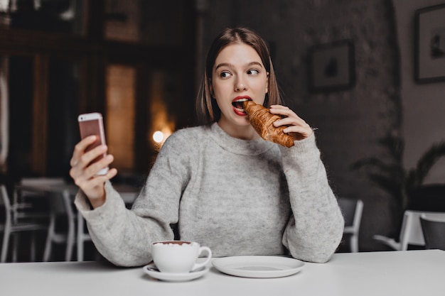 Jeune fille aux cheveux courts en sweat-shirt gris prend selfie en mordant un croissant croquant dans un café lumineux.