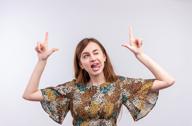 Jeune fille aux cheveux longs portant une robe colorée pointant l'index vers le haut qui sort la langue et un clin de œil