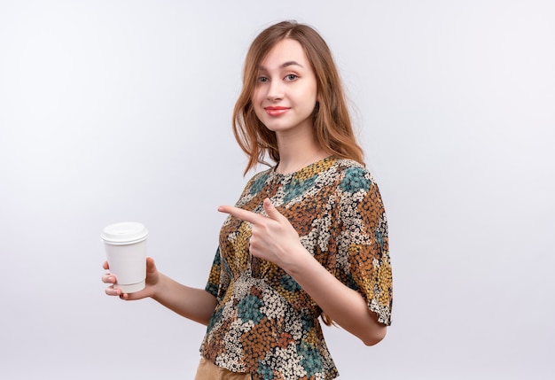 Jeune fille aux cheveux longs portant une robe colorée tenant une tasse de café pointant avec le doigt dessus