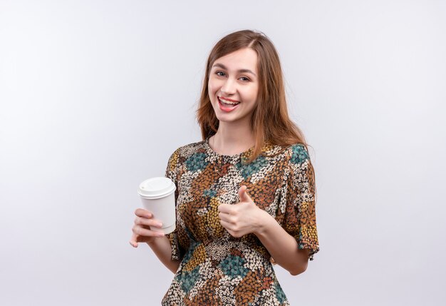 Jeune fille aux cheveux longs portant une robe colorée tenant une tasse de café souriant joyeusement montrant les pouces vers le haut