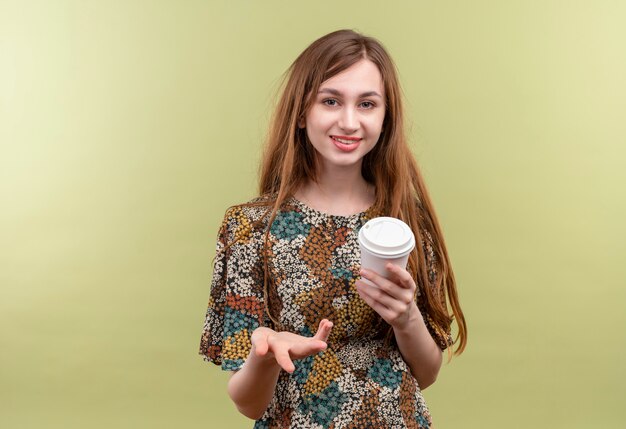 Jeune fille aux cheveux longs portant une robe colorée tenant une tasse de café en souriant regardant la caméra en levant la main comme question