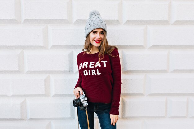 Jeune fille aux cheveux longs en pull marsala sur mur gris. Elle porte un bonnet tricoté, tient la caméra et sourit.