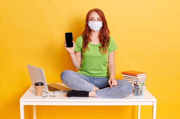 Jeune fille aux cheveux rouges en masque respiratoire tenant un téléphone intelligent avec écran blanc, recommander un nouvel appareil, mise en quarantaine covid d'apprentissage à distance, assis sur une table avec des livres, ordinateur portable, stylos.