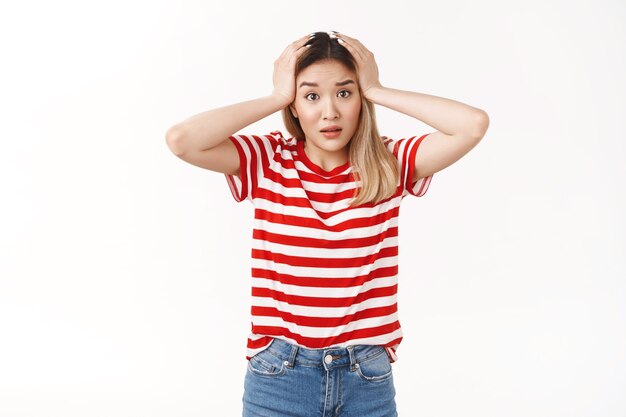 Une jeune fille blonde asiatique perplexe s'empare de la tête en train de paniquer, confus, qu'est-ce que l'appareil photo a l'air inquiet face à une situation terrible, réagissant nerveusement aux mauvaises nouvelles, haletant anxieux, debout sur un mur blanc