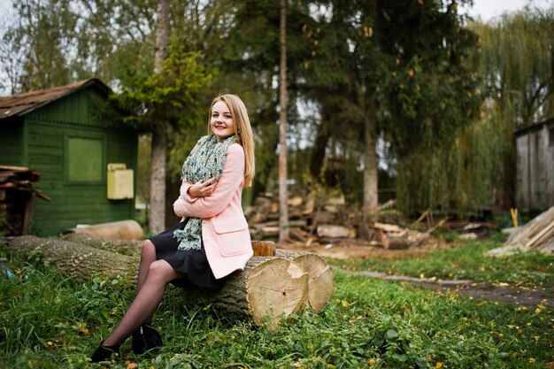 Jeune fille blonde au manteau rose posé sur le parc d'automne assis sur un arbre coupé contre la maison du forestier