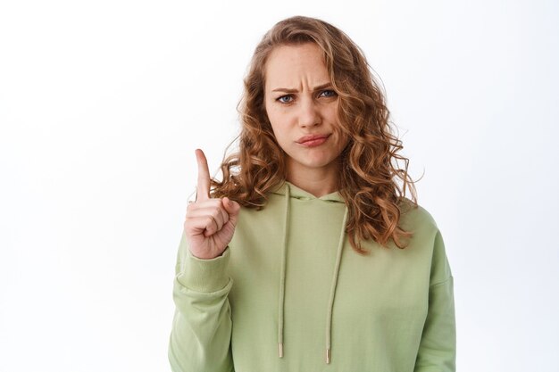 Jeune fille blonde bouleversée, fronçant les sourcils et ayant l'air déçue, pointant le doigt vers le haut du logo, jugeant une mauvaise chose, debout dans un sweat à capuche vert contre un mur blanc