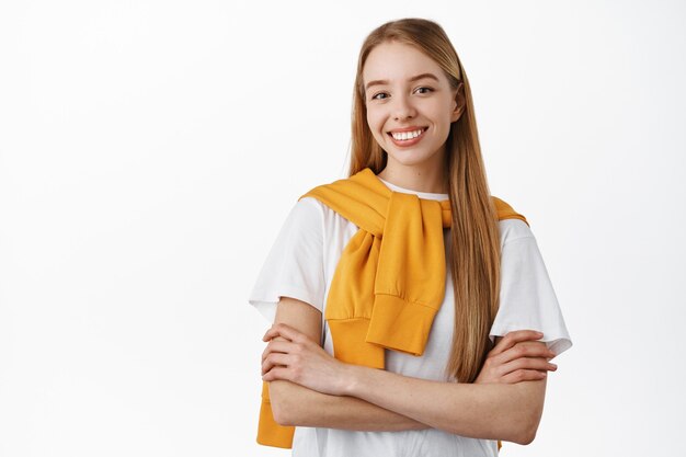 Jeune fille blonde confiante avec de longs cheveux naturels, les bras croisés sur la poitrine et souriante déterminée, debout comme une professionnelle, debout sur un mur blanc