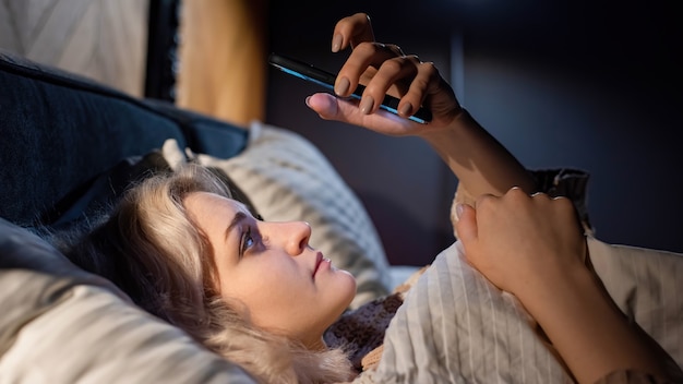 Jeune fille blonde est sur son smartphone dans le lit. Essayer de s'endormir. Dépendance aux médias sociaux