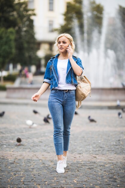 Jeune fille blonde femme parle téléphone sur fontain carré streetwalk habillé en suite de jeans bleu avec sac sur son épaule en journée ensoleillée