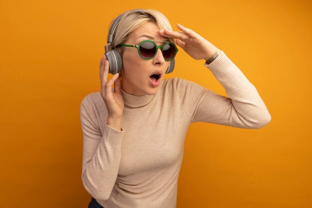 Jeune fille blonde impressionnée portant des lunettes de soleil et des écouteurs touchant des écouteurs mettant la main sur le front regardant le côté à distance