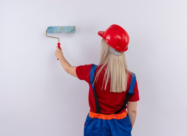 Jeune fille blonde ingénieur constructeur en peinture uniforme mur avec rouleau à peinture debout en vue de derrière sur mur blanc isolé