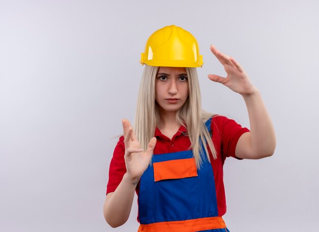 Jeune fille blonde ingénieur constructeur en uniforme montrant la forme de quelque chose à la recherche sur un mur blanc isolé avec copie espace