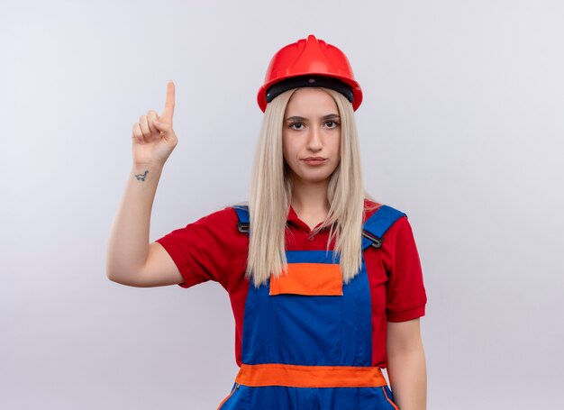 Jeune fille blonde ingénieur constructeur en uniforme pointant vers le haut et à la recherche sur le mur blanc isolé