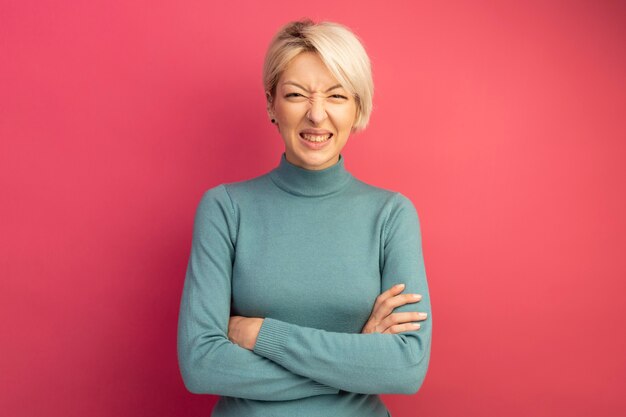 Jeune fille blonde irritée debout avec une posture fermée à la recherche d'isolement sur un mur rose avec espace de copie