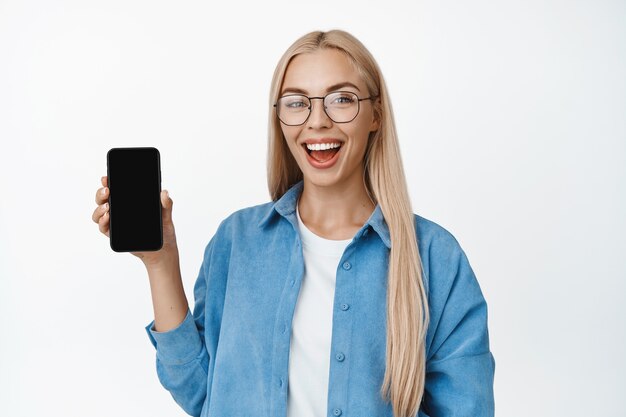 Jeune fille blonde à lunettes montrant l'écran du téléphone portable et souriant, debout dans des vêtements décontractés, démontrant l'application smartphone sur blanc.