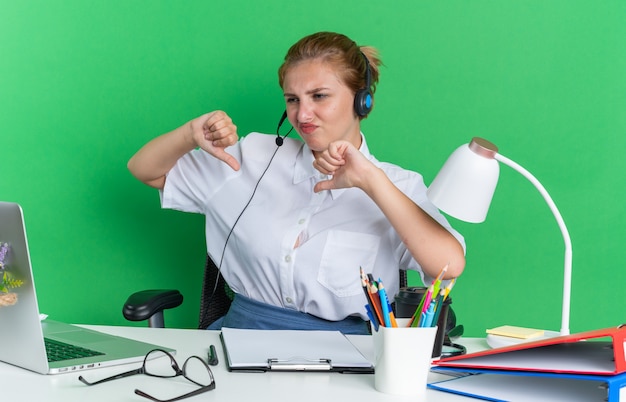 Jeune fille blonde mécontente du centre d'appels portant un casque assis au bureau avec des outils de travail en regardant un ordinateur portable montrant les pouces vers le bas