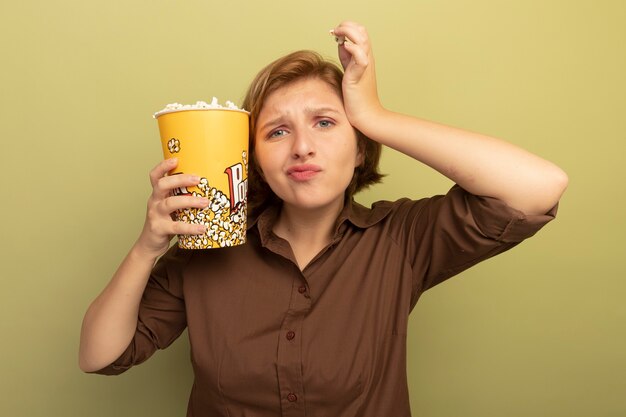 Jeune fille blonde mécontente tenant un seau de pop-corn près de la tête et un morceau de pop-corn touchant la tête en regardant la caméra isolée sur fond vert olive