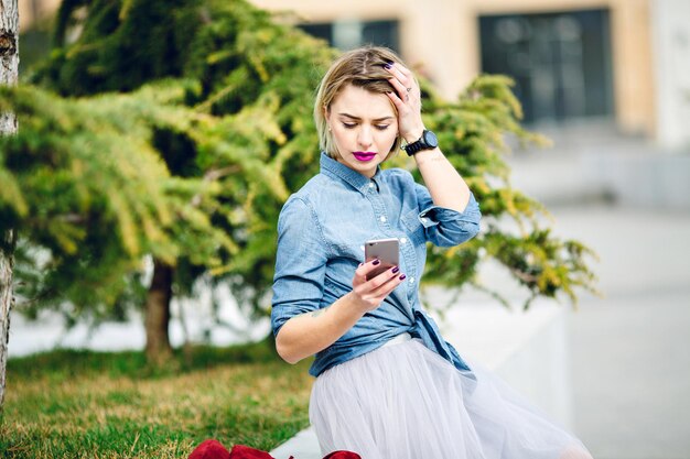 Jeune fille blonde mignonne aux cheveux courts et aux lèvres rose vif, assise dans un parc et lisant un message sur son smartphone, vêtue d'une chemise bleu denim et d'une jupe en tulle gris.