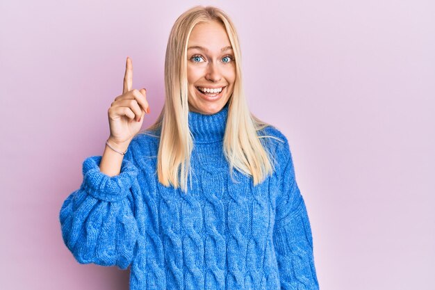 Jeune fille blonde portant un pull d'hiver en laine pointant le doigt vers le haut avec une idée réussie. excité et heureux. numéro un.