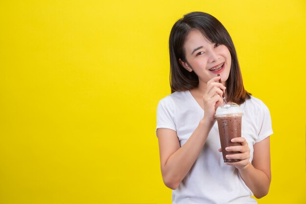 La jeune fille boit de l'eau froide dans du cacao dans un verre en plastique transparent sur un fond jaune.