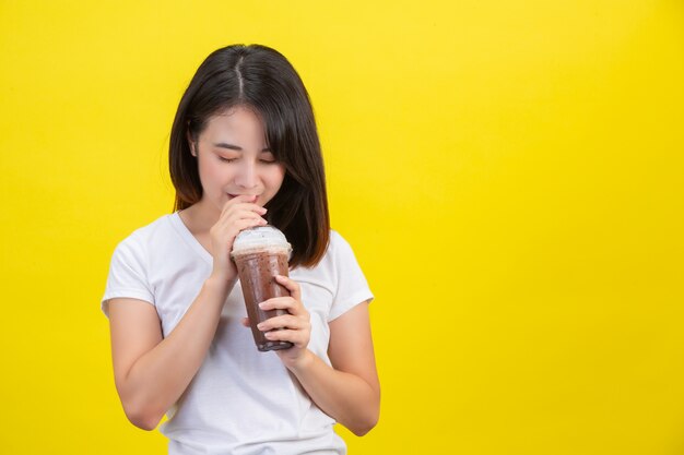 La jeune fille boit de l'eau froide dans du cacao dans un verre en plastique transparent sur un fond jaune.