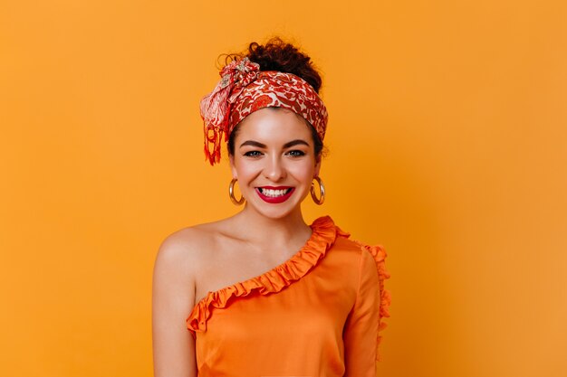 Jeune fille de bonne humeur sourit sur l'espace orange. Élégante dame aux cheveux noirs en chemisier orange et écharpe sur la tête se penche sur la caméra.