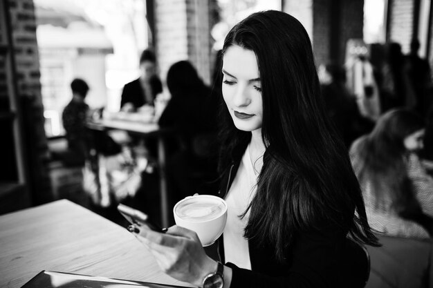 Jeune fille brune assise sur un café avec une tasse de cappuccino travaillant avec un ordinateur portable rouge et regardant un téléphone portable