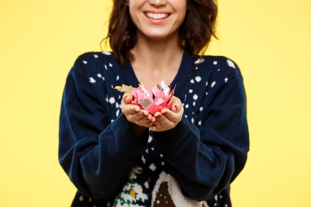 Jeune fille brune belle joyeuse en pull tricoté confortable souriant avec une poignée de confettis sur mur jaune
