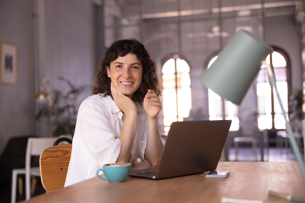 Jeune fille brune caucasienne positive regardant la caméra assise à table avec un ordinateur portable à l'intérieur