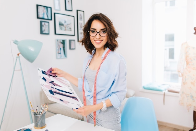 Une jeune fille brune dans une robe grise et une chemise bleue se tient près de la table dans un atelier de studio. Elle a des échantillons et des croquis dans ses mains. Elle sourit à la caméra.