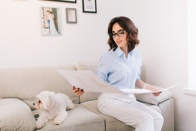 Une jeune fille brune est assise sur le canapé en studio. Elle porte une chemise bleue et un pantalon blanc. Elle regarde les croquis qui sont entre ses deux mains.