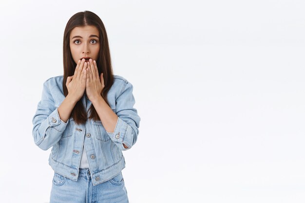 Une jeune fille brune innocente et timide choquée a l'air peu sûre et inquiète, légèrement effrayée, haletant la bouche avec la paume et regarde la caméra nerveuse, debout anxieuse et préoccupée sur fond blanc