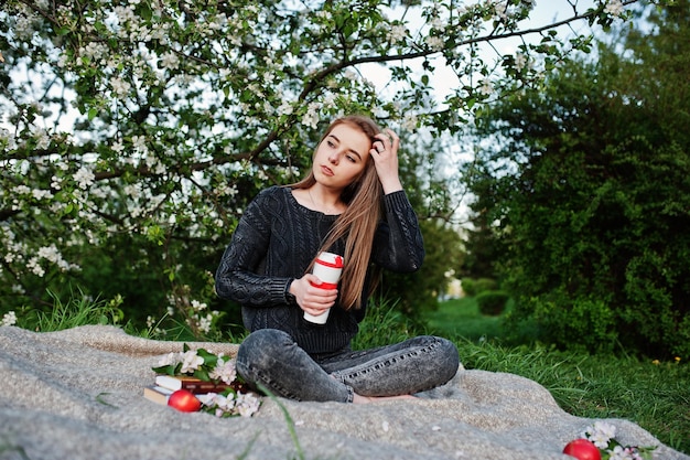 Jeune fille brune en jeans assis sur un plaid contre un arbre de fleurs de printemps et tenant un thermos de thé à la main
