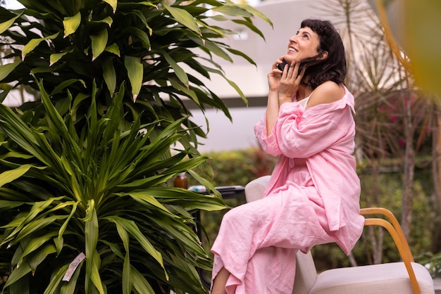 Photo gratuite jeune fille brune à la peau claire dans des vêtements décontractés lève les yeux à l'aide d'un téléphone assis dans un jardin tropical pendant la journée concept de style de vie