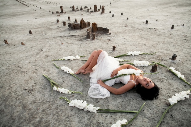 Photo gratuite jeune fille brune en robe blanche aux yeux fermés est allongée sur le littoral entouré de glaïeuls blancs