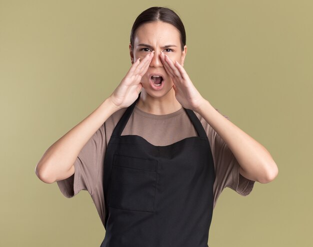 Une jeune fille brune sérieuse de barbier en uniforme tient les mains près de la bouche appelant quelqu'un isolé sur un mur vert olive avec espace de copie