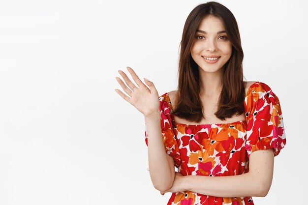 Jeune fille brune souriante agitant la main pour dire bonjour portant une robe à fleurs d'été debout sur fond blanc