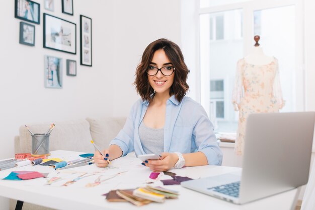 Une jeune fille brune souriante dans une chemise bleue est assise à la table en studio. Elle dessine avec des crayons. Elle sourit à la caméra.