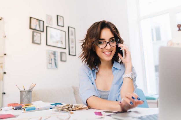 Une jeune fille brune souriante dans une chemise bleue travaille dans l'atelier. Elle parle au téléphone et tape sur ordinateur. Elle sourit à la caméra.