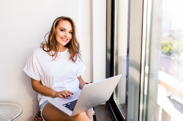 Photo gratuite jeune fille brune souriante est assise sur une chaise moderne près de la fenêtre dans une pièce confortable et lumineuse à la maison. elle travaille sur ordinateur portable dans une atmosphère relaxante