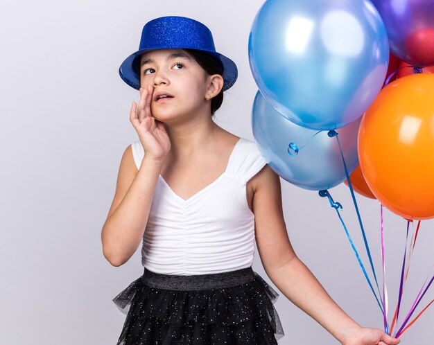 jeune fille caucasienne choquée avec un chapeau de fête bleu tenant des ballons à l'hélium et levant isolé sur un mur blanc avec espace de copie